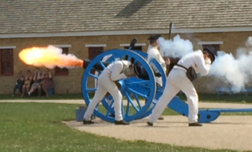 Two early American soldiers cover their ears as fire leaves the cannon's barrel
