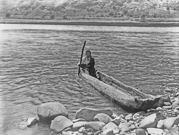 Nez Perce man with pole maneuvering dugout canoe to rocky shore